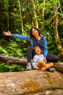 Cheerful mother and her son are sitting on a large fallen log in cathedral grove forest on
