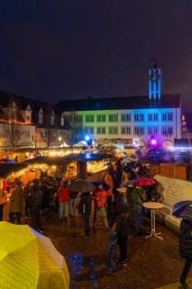 Colourfully lit Christmas market with people and umbrellas in a nighttime urban landscape,