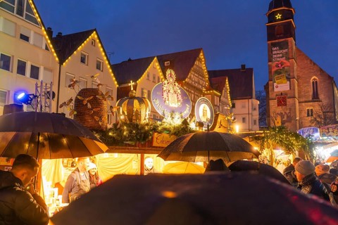 Crowd of people with umbrellas at an illuminated Christmas market in front of a church at night,