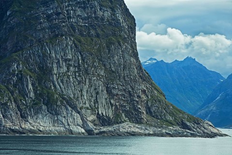A massive rock juts out of the calm water, surrounded by a dramatic mountain backdrop under a