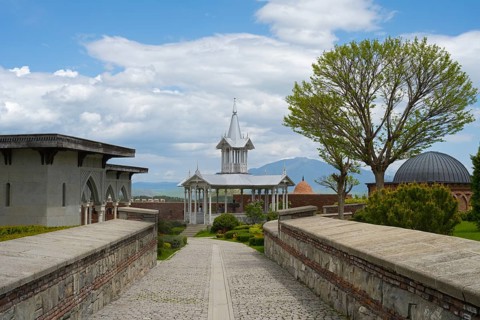 Architectural structure with tower and trees under a cloudy sky along a paved path, Rabati