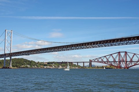 Two distinctive bridges over water with a small sailboat in the foreground, railway bridge and road