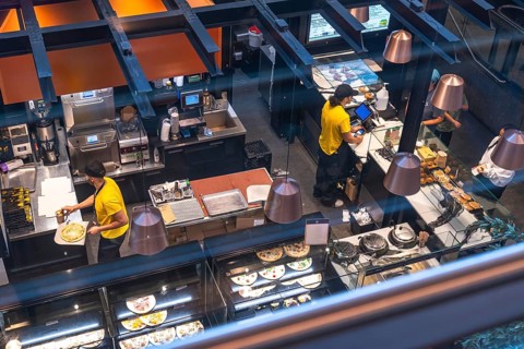 Chefs working in a busy airport restaurant kitchen prepare various dishes, including pizza and