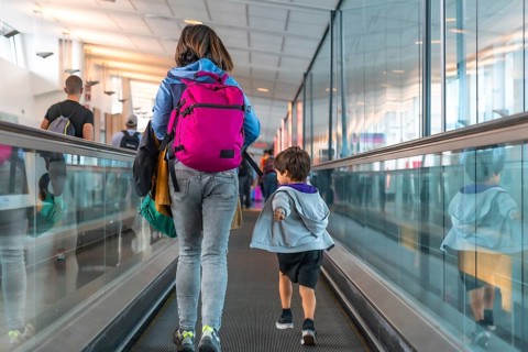 Mother and son are walking on a moving walkway at the airport, carrying backpacks and luggage as