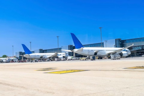 Two large passenger airplanes are parked at the gate of a modern airport on a sunny day, undergoing