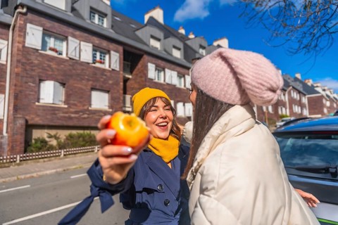 Two young women laughing and sharing a healthy snack on a sunny urban street, bundled in coats and