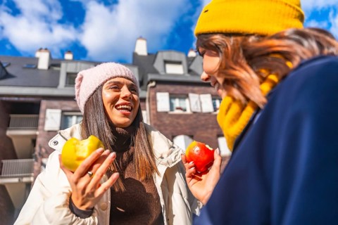 Two cheerful women friends enjoying a healthy apple snack and talking outdoors on a sunny winter