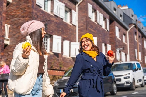 Smiling young women friends walking and talking on a city street, enjoying a healthy snack of