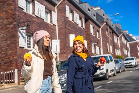 Two cheerful young women enjoying a healthy snack of apples while walking and smiling on a city