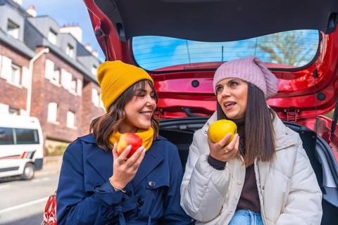 Two cheerful women sit in a car trunk on a winter road trip, sharing apples, laughing and talking
