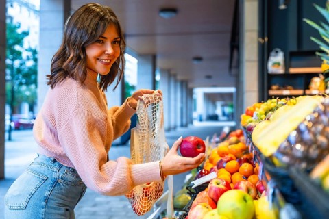 Young woman smiling while adding fresh red apples to her reusable mesh bag at an outdoor or street