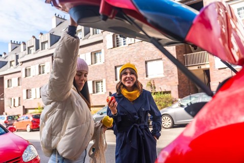 Two cheerful young women are holding apples and smiling, standing together by the open trunk of a