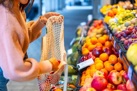 Woman selecting organic apples and oranges, placing them into a sustainable net shopping bag while