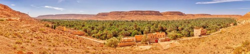 A wide panorama with an oasis, many palm trees and buildings against a mountain backdrop, Ziz