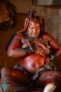 Elderly Himba woman rubbing herself with red paste for skin care, in a hut, traditional Himba