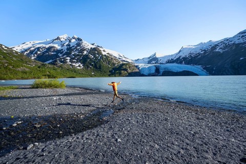 Young man jumping on a pebble beach on a glacial lake, Snowy Mountains and Portage Glacier on