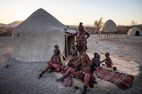 Married Himba woman with their babies in their arms sitting in front of the first woman's hut, in