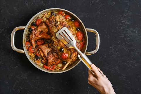 A hand uses a kitchen tongs to lift a tender rabbit leg from a pan filled with roasted tomatoes and