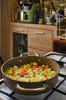 Chopped leeks and carrots sizzle in a large pot on a stovetop. The kitchen features wooden