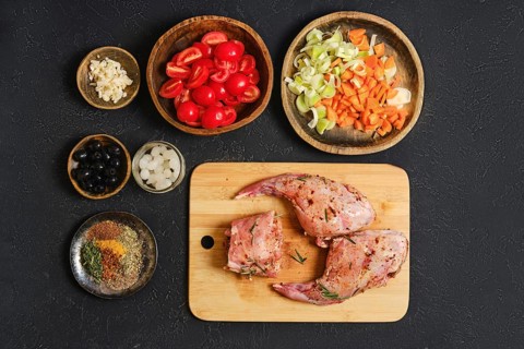 A rabbit's hind legs and a saddle are placed on a wooden cutting board, surrounded by fresh