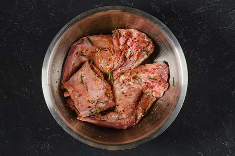Two raw hind leg and saddle of rabbit is marinating with herbs and spices in a metal bowl. It is