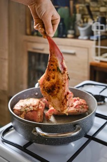 A person holds a marinated rabbit hind leg above a pot with other meat cooking inside. The kitchen