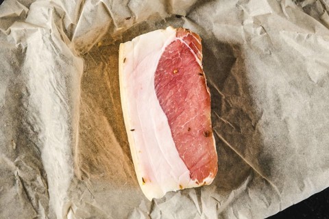 A slice of cured meat lies on a piece of parchment paper in a kitchen. The meat features a mix of