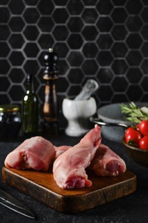 Rabbit hind legs sit on a wooden cutting board, surrounded by herbs, tomatoes, and kitchen tools.