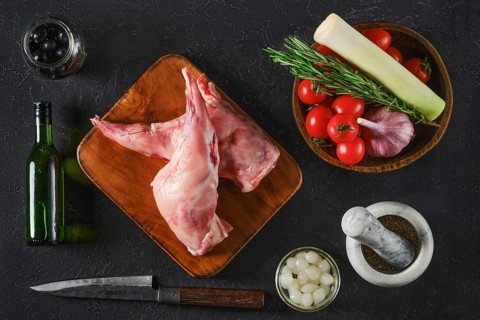 Top view of two raw rabbit hind legs are on a wooden board surrounded by fresh vegetables, herbs,