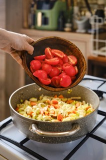 A person is adding chopped red tomatoes from a wooden bowl into a pot that is already filled with