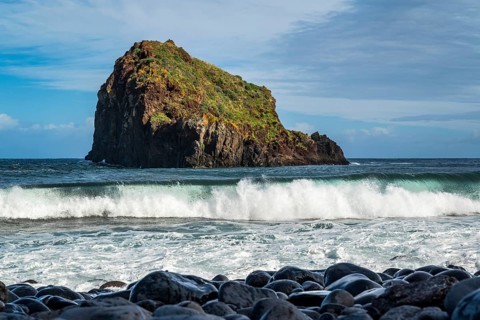 View over black rocks to the surf and a rolling wave off the island of Ilhéu da Ribeira da Janela,