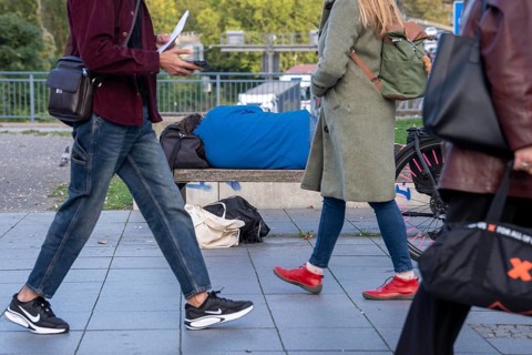 Man sleeping on a bench, travelling bag serves as pillow, in the city centre, passers-by walk by