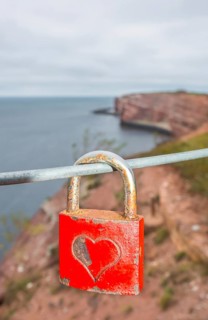 Red padlock with engraved heart symbol on a wire rope in front of the steep coast, red cliff of