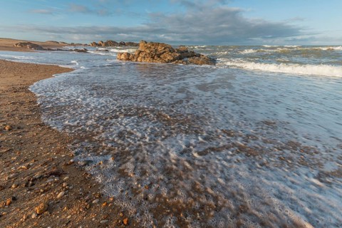A wide sandy beach stretches out with scattered pebbles and beach shells. Gentle waves crash