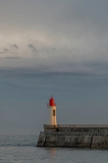 A lighthouse stands on a pier and casts its light over calm waters at dusk. The sky is filled with