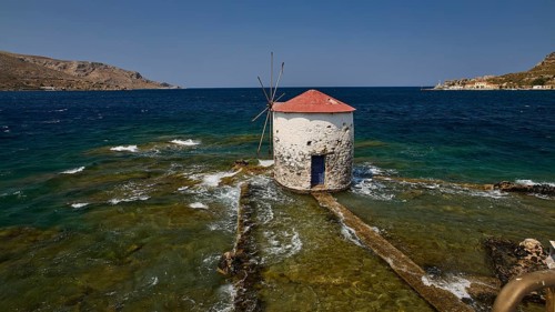 Historic windmill stands in the turquoise sea, surrounded by waves and a clear sky, Agia Marina,