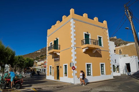 Mediterranean-style corner building with balconies and passers-by under a clear blue sky, Agia