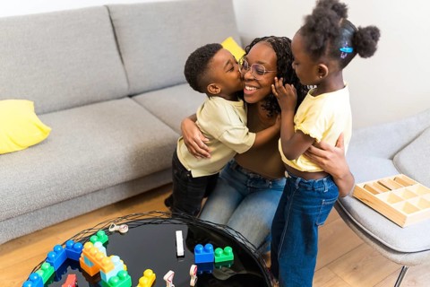 Cheerful mother enjoying time with her two loving children, embracing and kissing in the living