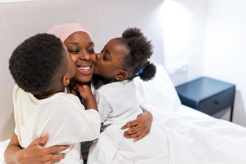 Two children kissing their smiling mother with cancer in bed, enjoying family time together