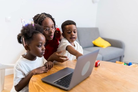 Young mother guiding her two children while using a laptop on a wooden table at home, fostering