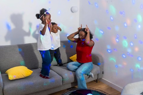 Mother making funny faces while kids dancing joyfully on the sofa, illuminated by colorful disco
