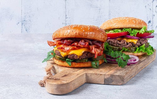 Two burgers with beef patty, fried bacon, cheddar cheese, tomatoes, on a light table
