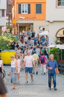 People stroll through a lively old town street, surrounded by shops and summery impressions, 950
