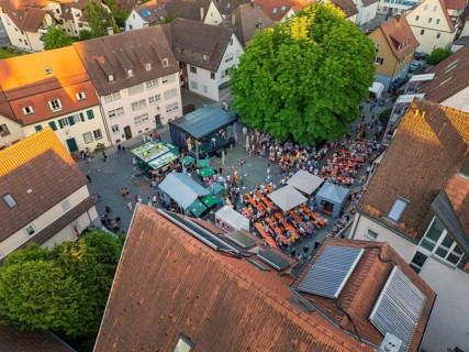 Aerial view of a lively town festival, surrounded by orange-coloured roofs and green trees in