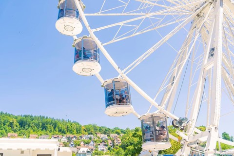 Ferris wheel with cabins in front of a blue sky, green trees and houses in the background, Calw,