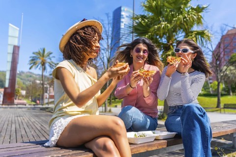 Three young women savoring delicious pizza while sitting on a bench in a vibrant city park,