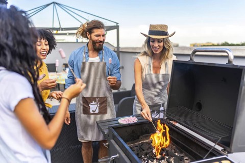 Happy friends wearing aprons are toasting marshmallows on a barbecue grill during a rooftop party
