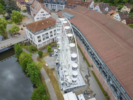 A large white Ferris wheel stands next to modern buildings with red roofs in an urban setting,