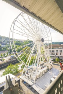 A Ferris wheel is set up in a town next to a river, surrounded by green landscape and buildings,