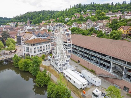 A Ferris wheel next to urban buildings embedded in a summer landscape with wooded hills, Ferris